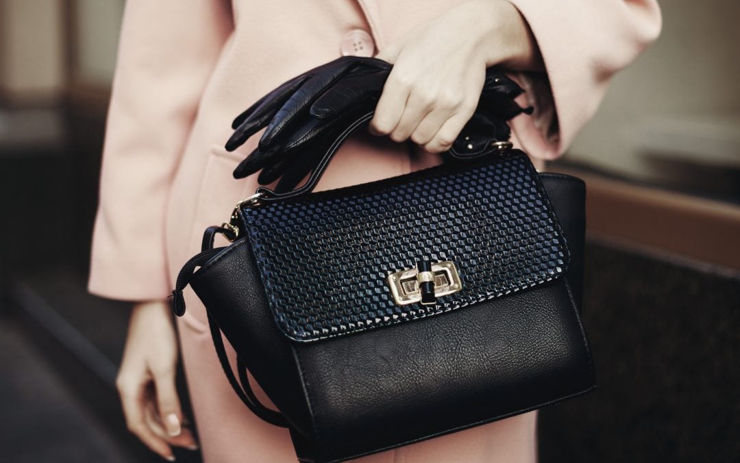 Eine sehr stilvolle Frau, die eine schwarze Handtasche und schwarze Handschuhe trägt, der Mantel ist lachsfarben. Man sieht nur die Hände und den Oberkörper