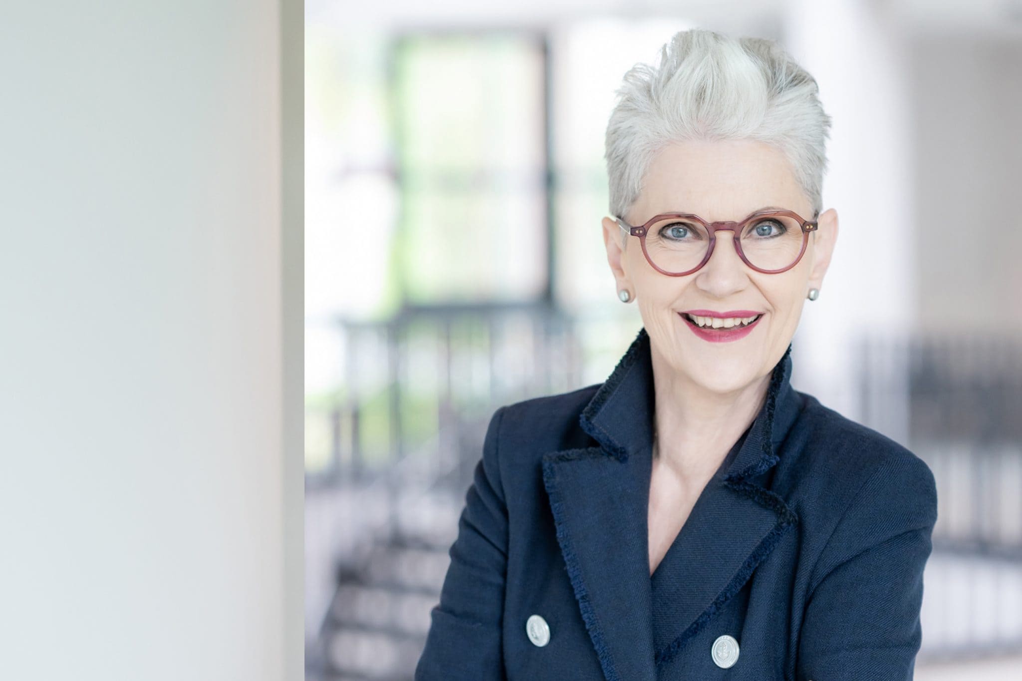 Elisabeth Motsch trägt eine rote Brille, hat weißes Haar und hat einen roten Lippenstift. Sie trägt einen dunkelblauen Blazer mit Silberknöpfen. Der Kragen ist aufgestellt.