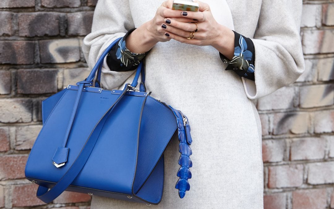 Ein Frau die sehr stilvoll und trendig angezogen ist. Sie trägt eine royalblaue Handtasche. Die Farbe der Handtasche findet sich in der schwarzgemusterten Bluse wieder. Der Mantel ist silbrig. Sie hat ein Handy in der Hand, wo sie gerade tippt. Man sieht sie nur bis zum Oberkörper