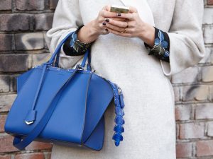 Ein Frau die sehr stilvoll und trendig angezogen ist. Sie trägt eine royalblaue Handtasche. Die Farbe der Handtasche findet sich in der schwarzgemusterten Bluse wieder. Der Mantel ist silbrig. Sie hat ein Handy in der Hand, wo sie gerade tippt. Man sieht sie nur bis zum Oberkörper